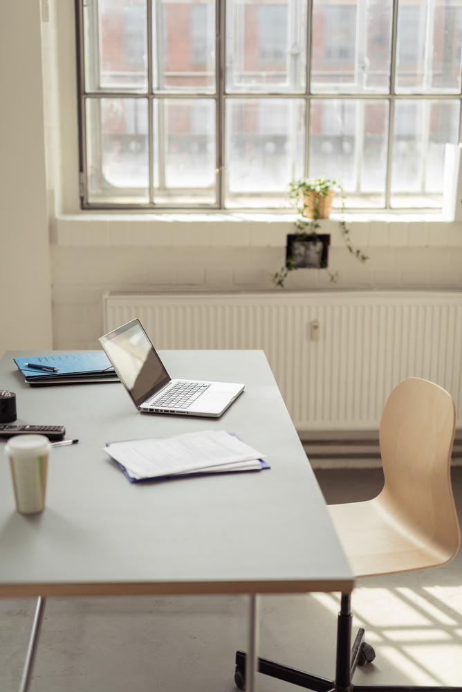 Laptop, documents and a paper coffee cup on a white desk in the interior of a modern bright office with large window, radiator and a wooden chair on wheels